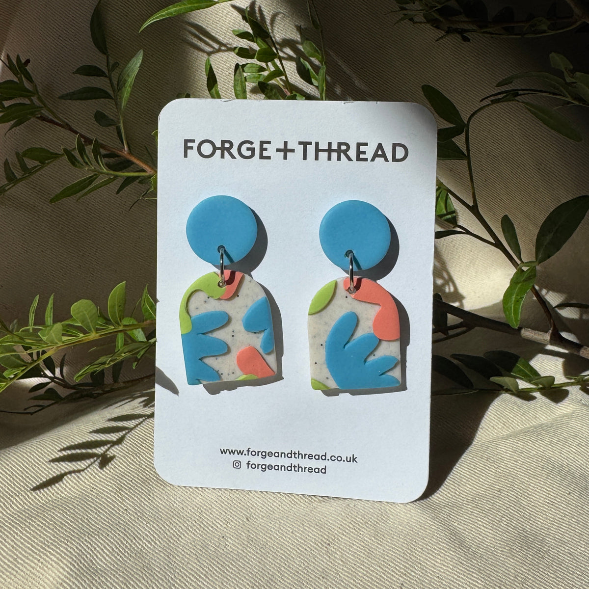A pair of colorful abstract earrings displayed on a white card labeled FORGE+THREAD, set against a draped beige fabric backdrop with green leaves.