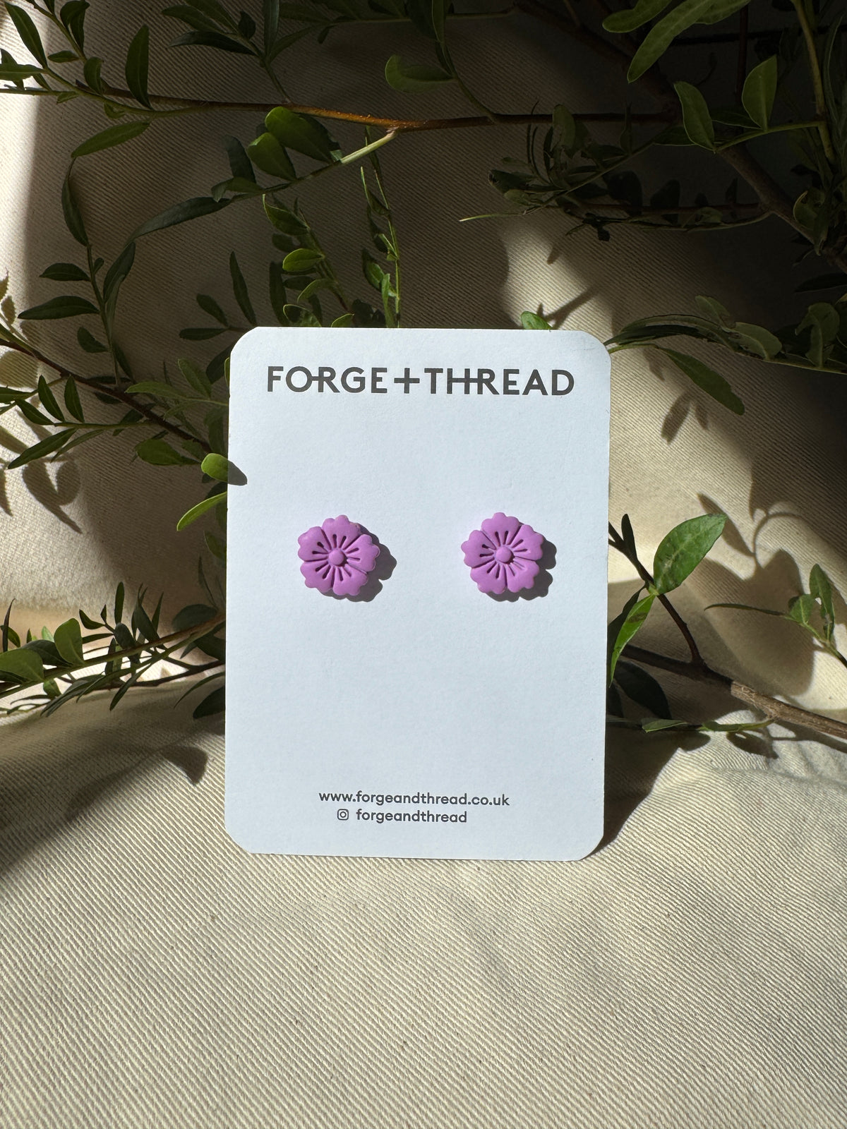 A pair of small purple flower-shaped stud earrings displayed on a white card labeled FORGE+THREAD, set against a beige fabric background with green leaves.