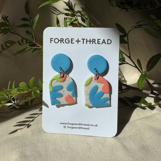 A pair of colorful abstract earrings displayed on a white card labeled FORGE+THREAD, set against a draped beige fabric backdrop with green leaves.