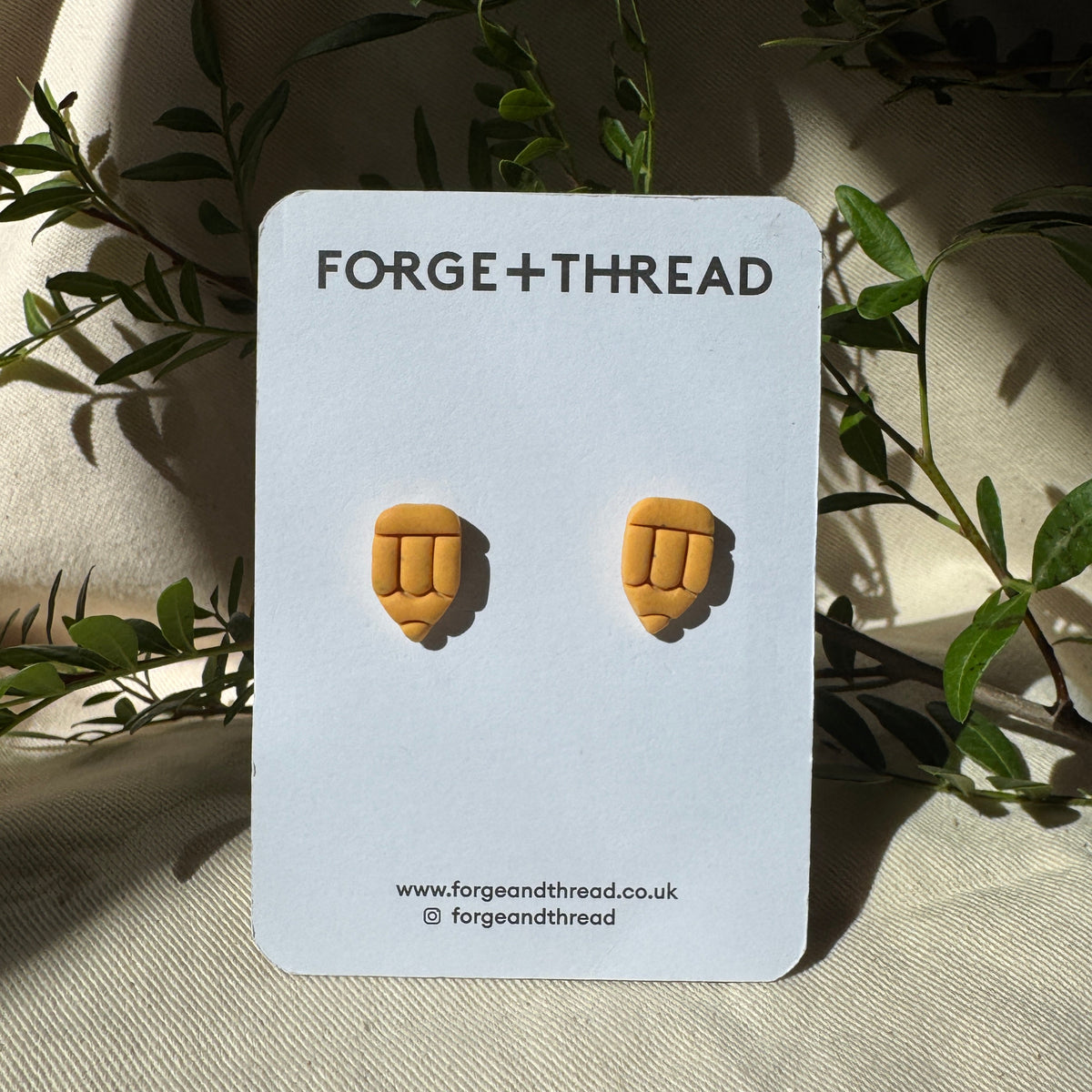 A pair of small yellow-orange stud earrings shaped like clenched fists, mounted on a white card labeled FORGE+THREAD, photographed against draped fabric with green leaves around it.