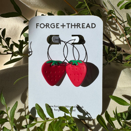 A pair of strawberry-shaped hoop earrings displayed on a white card labeled FORGE+THREAD, styled with green leaves on a beige fabric background.
