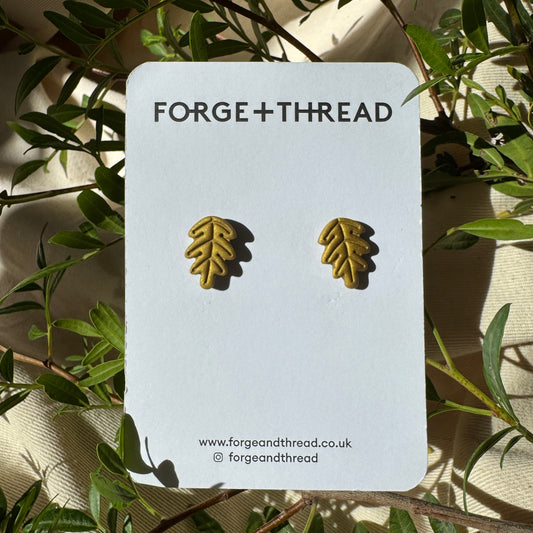 A pair of small gold leaf stud earrings displayed on a white Forge+Thread backing card, surrounded by green leaves and branches.