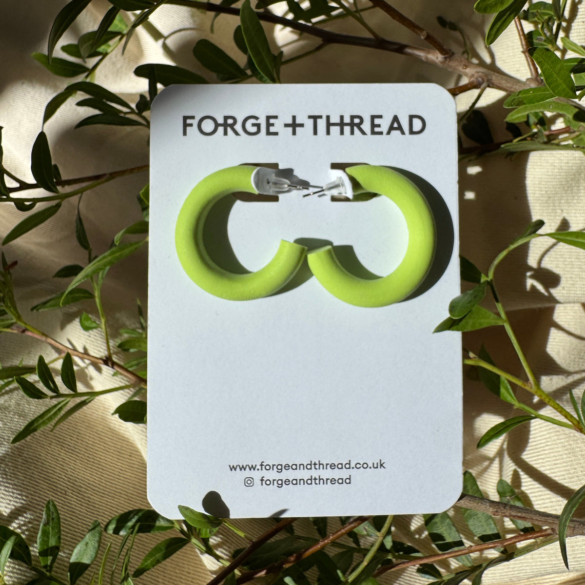 A pair of lime green hoop earrings displayed on a white card labeled 'FORGE+THREAD', placed among green leaves and draped fabric.
