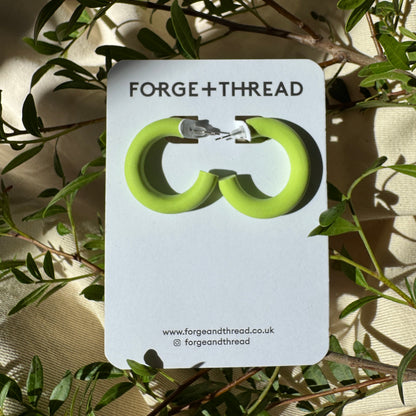 A pair of lime green hoop earrings displayed on a white card labeled 'FORGE+THREAD', placed among green leaves and draped fabric.