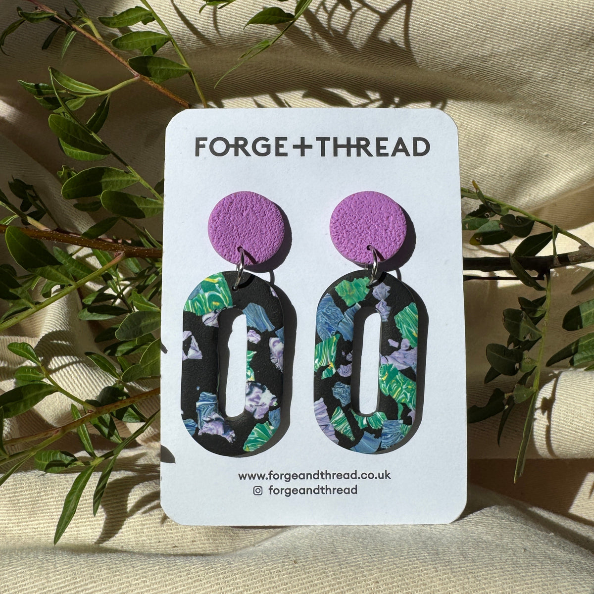 A pair of statement earrings displayed on a white card labeled FORGE+THREAD, placed on a beige fabric surface with green leaves around it. The earrings have a textured purple circular top and a black base with a green, blue, and white floral pattern.