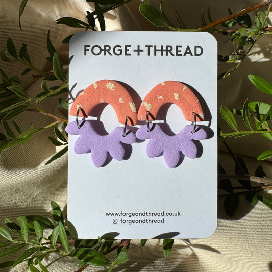 A pair of handmade arch-shaped earrings by Forge + Thread, featuring a coral orange top half with tiny white dots and a lavender purple bottom half, displayed on a white backing card surrounded by green leaves on a beige fabric background.