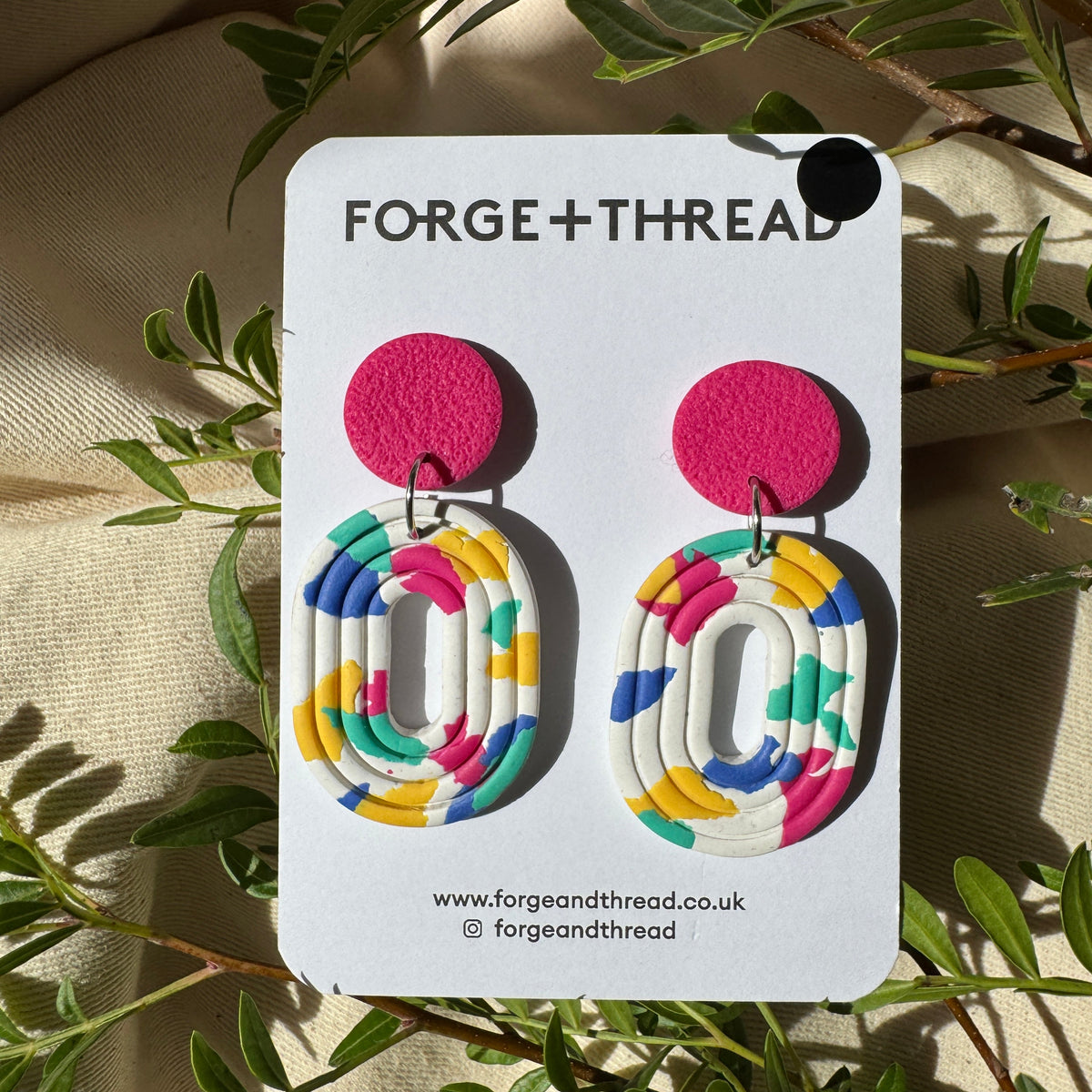 A pair of colorful geometric dangle earrings displayed on a white card labeled FORGE+THREAD, placed on a beige fabric surface with green leaves around it.