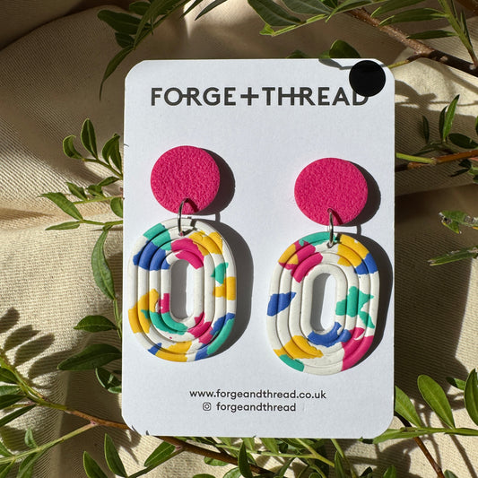 A pair of colorful geometric dangle earrings displayed on a white card labeled FORGE+THREAD, placed on a beige fabric surface with green leaves around it.