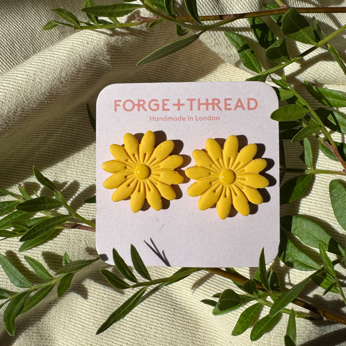 A pair of yellow daisy stud earrings on a white display card reading 'FORGE+THREAD Handmade in London', styled with green leaves on a beige fabric background.