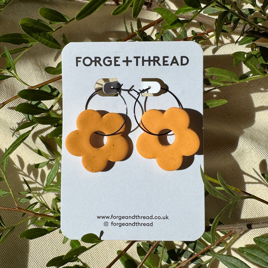 A pair of orange flower-shaped hoop earrings displayed on a white Forge+Thread card, photographed outdoors among green leaves and natural sunlight.