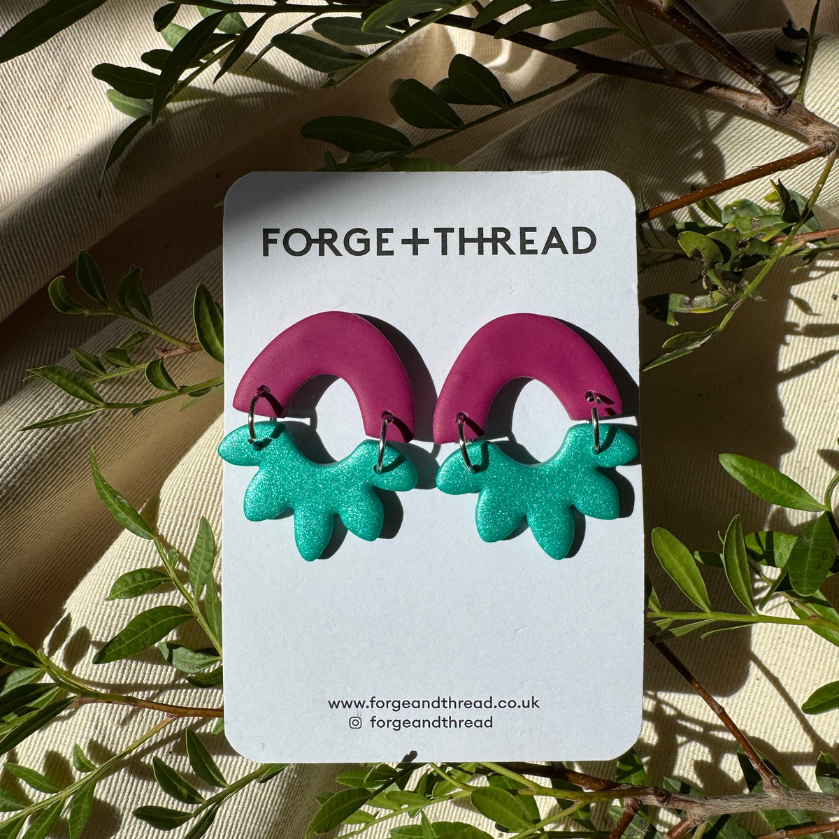 A pair of handmade arch-shaped earrings by Forge+Thread, featuring a magenta top half and a teal bottom half, displayed on a white brand card surrounded by green leaves and beige fabric.