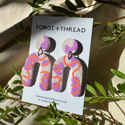 A pair of sculptural earrings displayed on a white card labeled FORGE+THREAD, placed on beige fabric with green leaves around it. The earrings have a lavender-purple base with an orange wavy line pattern and a small silver hoop connector.