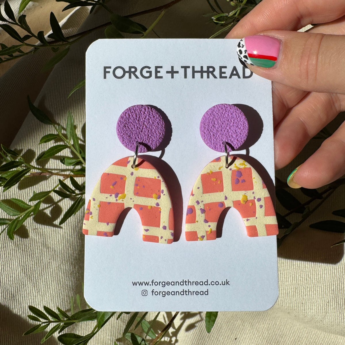 A hand holds a white card displaying a pair of statement earrings. The earrings have purple textured circular tops and arch-shaped lower halves with a pink, orange, and yellow checkered pattern.