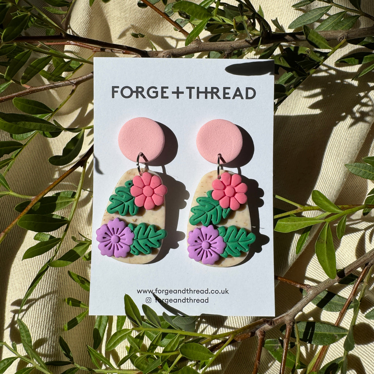 A pair of handmade polymer clay earrings displayed on a white card labeled 'FORGE+THREAD', placed on a cream fabric background with green leaves and branches around it. The earrings feature pink flower tops, beige stone-like bodies, green leaves, and pink and purple flowers.