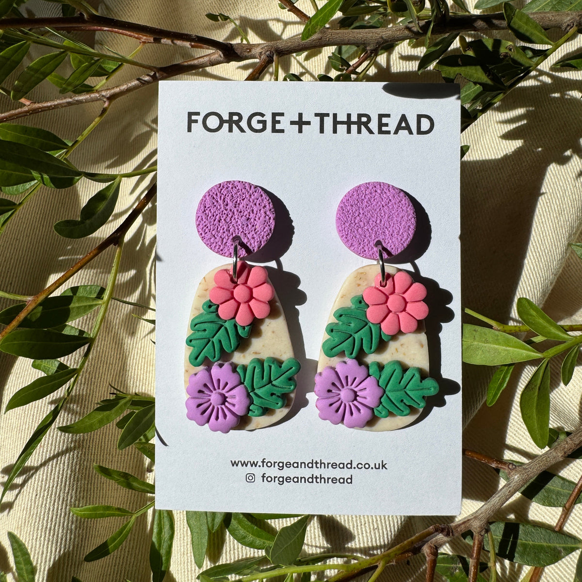 A pair of handmade dangle earrings displayed on a white card labeled 'FORGE+THREAD'. The earrings feature a purple textured circular top, a beige irregular lower section with pink flowers and green leaves, and a smaller purple flower at the bottom.