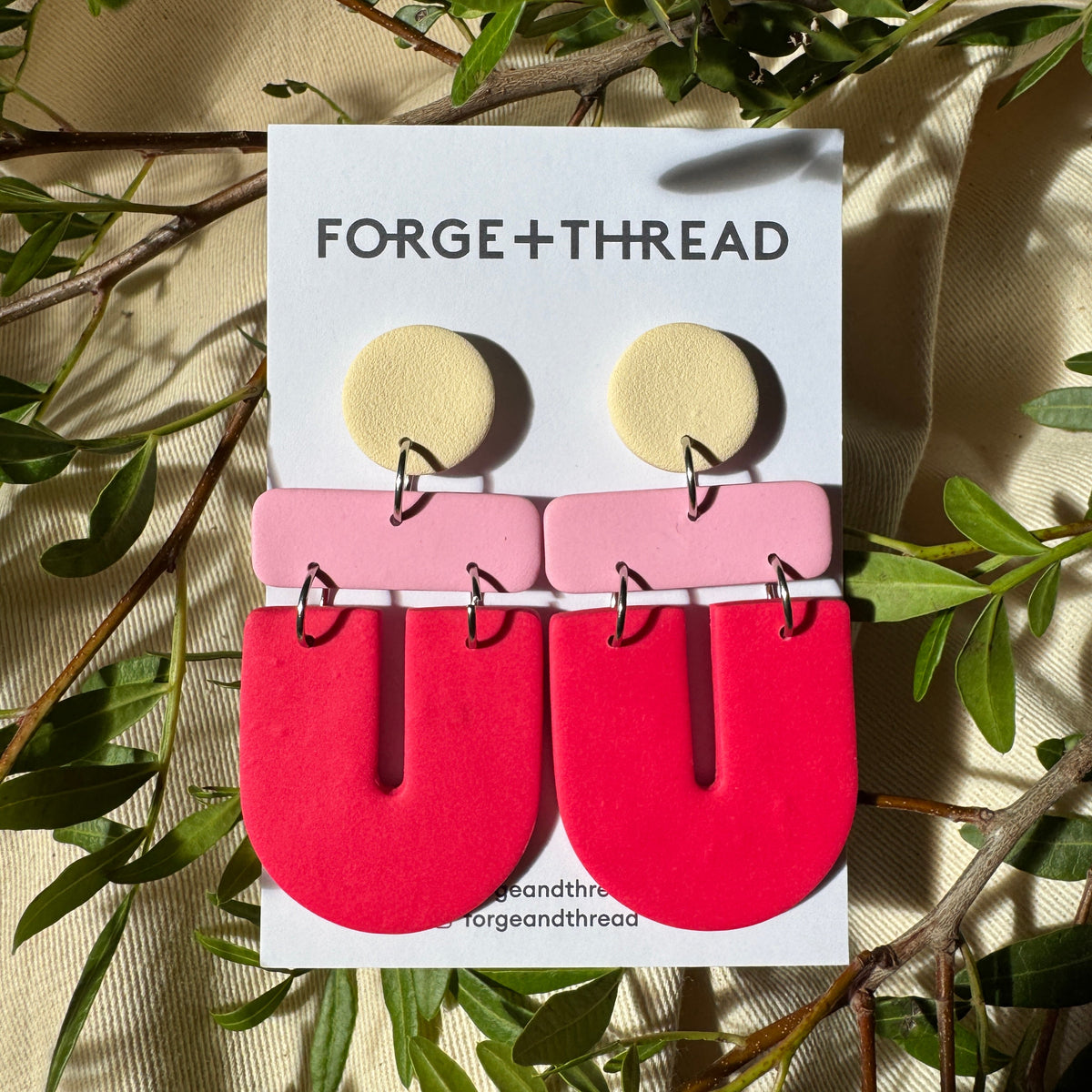 A pair of geometric statement earrings by Forge+Thread, featuring a pale yellow circular stud, a light pink rectangular bar, and a large bright red U-shaped drop, displayed on a white brand card surrounded by green leaves.
