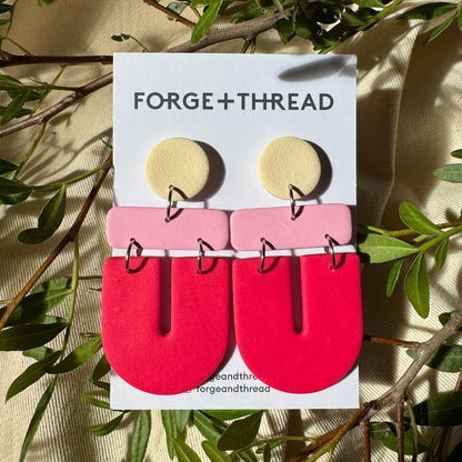 A pair of geometric statement earrings by Forge+Thread, featuring a pale yellow circular stud, a light pink rectangular bar, and a large bright red U-shaped drop, displayed on a white brand card surrounded by green leaves.