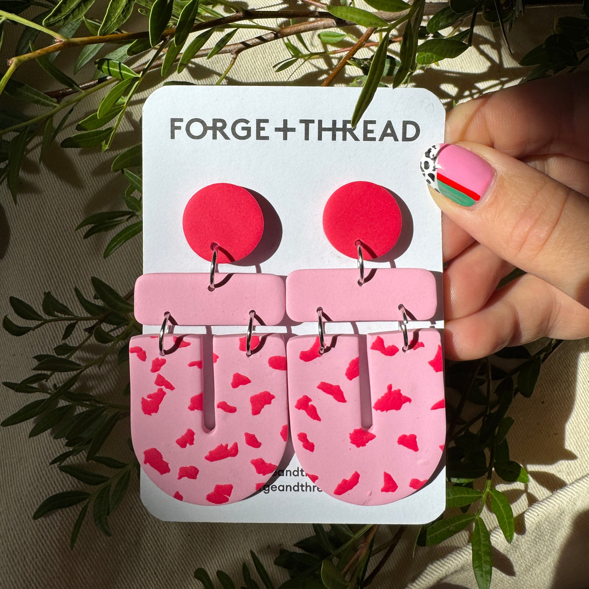 A pair of pink and red statement earrings on a white display card labeled 'FORGE+THREAD', held by a hand with pink nail art against a background of green leaves and beige fabric.