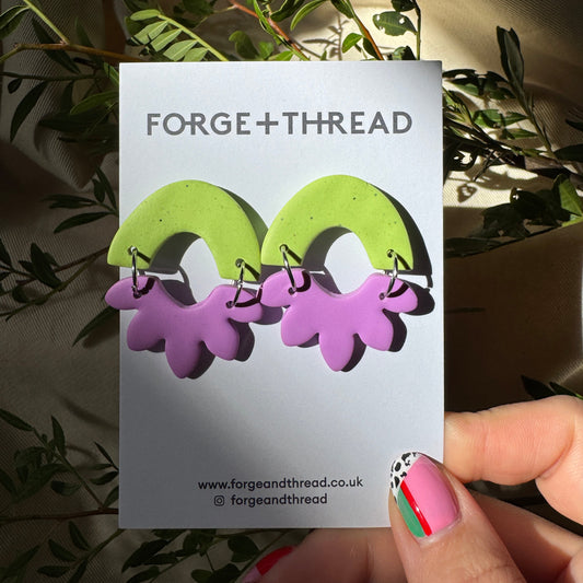 A pair of handmade polymer clay earrings by Forge+Thread, featuring lime green arches with dangling light purple flower charms, displayed on a branded white backing card held by a hand.