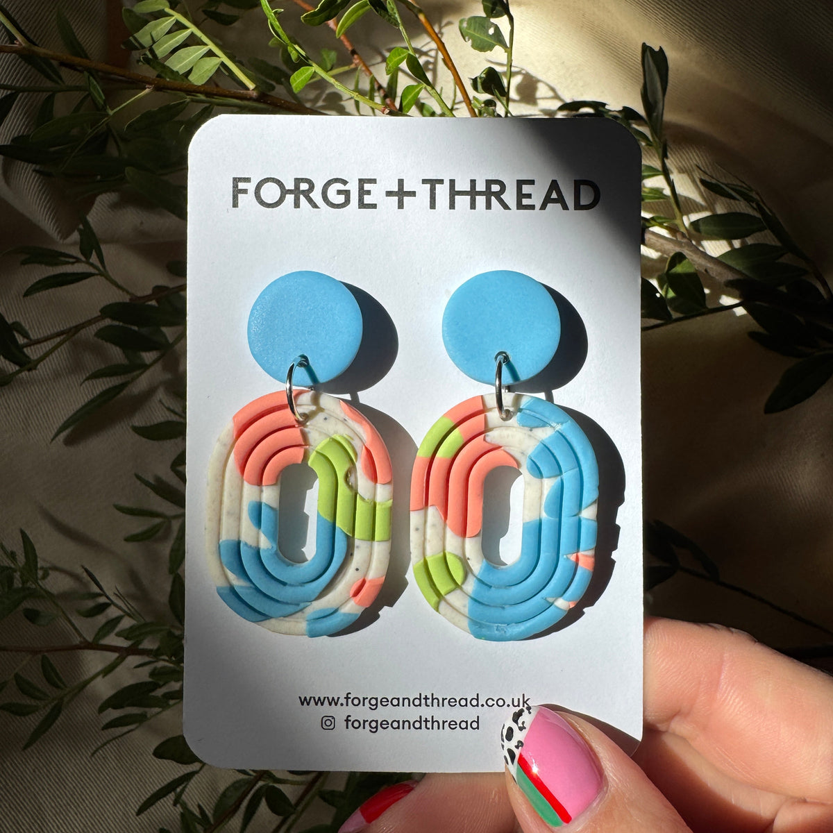A hand holds a white display card featuring a pair of colorful geometric drop earrings. The earrings have bright blue circular studs and wavy, layered dangle charms in pastel blue, pink, green, and white.