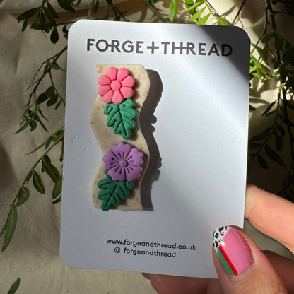 A small handmade accessory on a white display card, held by a hand with pink nail art. The accessory features pink and purple flowers with green leaves on a beige background.