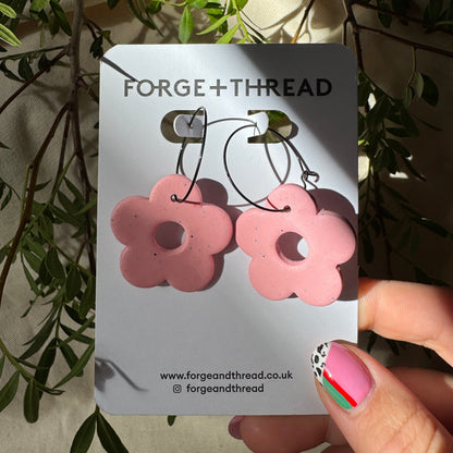 A pair of pink flower-shaped hoop earrings on a white Forge+Thread display card, held by a hand with decorated fingernails against a background of olive branches and cream fabric.