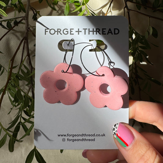 A pair of pink flower-shaped hoop earrings on a white Forge+Thread display card, held by a hand with decorated fingernails against a background of olive branches and cream fabric.
