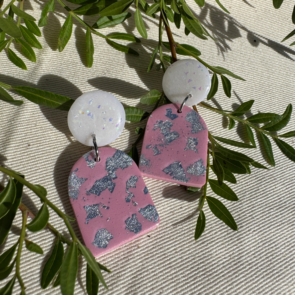 A pair of handmade drop earrings with round white iridescent studs and shield-shaped pink dangles covered in silver glitter, styled on a beige textured fabric with green leaves.