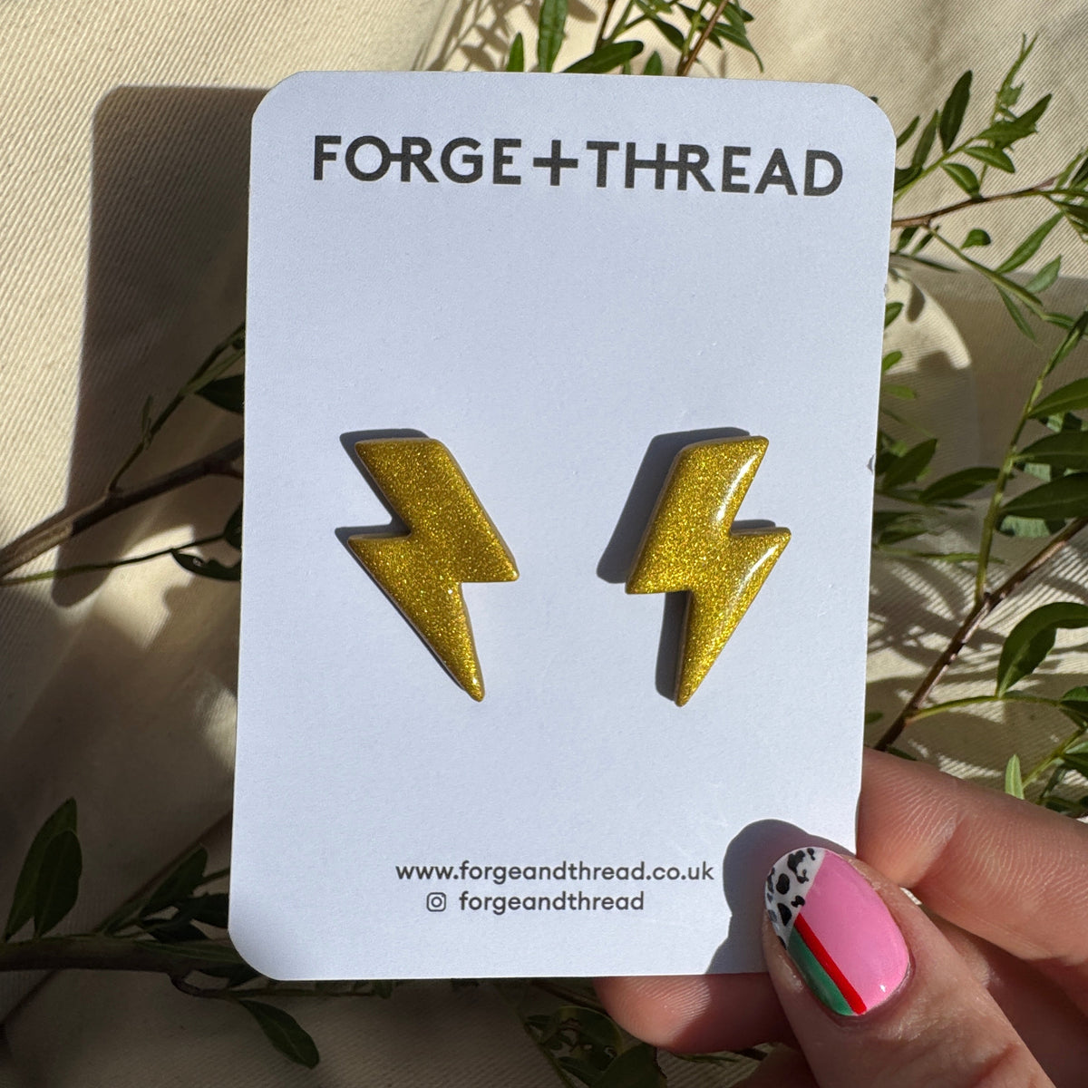 A pair of gold glitter lightning bolt stud earrings displayed on a white card labeled FORGE+THREAD, held by a hand with colorful nail art against a beige fabric background with green leaves.