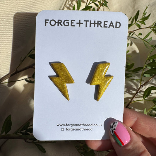 A pair of gold glitter lightning bolt stud earrings displayed on a white card labeled FORGE+THREAD, held by a hand with colorful nail art against a beige fabric background with green leaves.