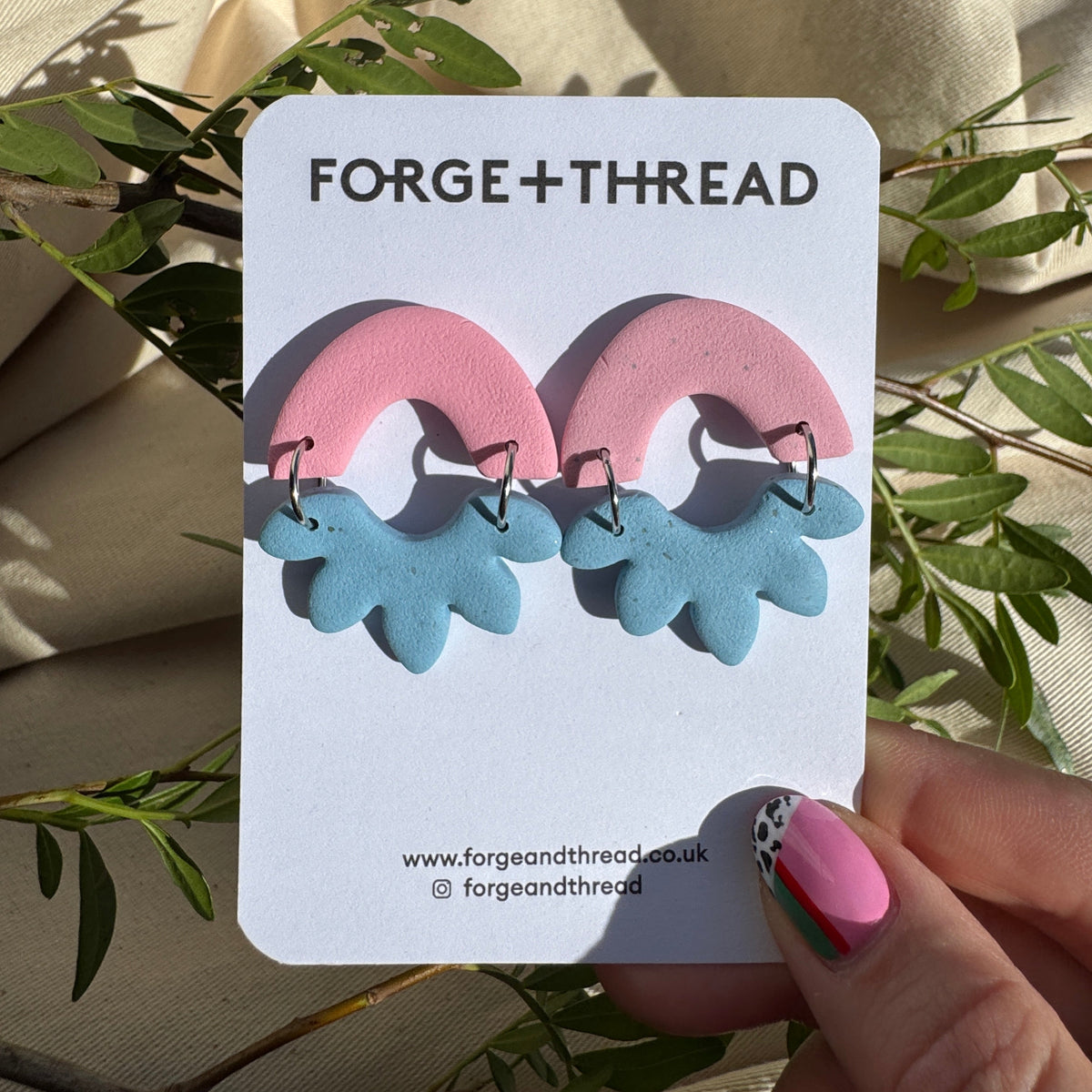 A pair of handmade polymer clay earrings by Forge + Thread, featuring pink arches with light blue leaf-like dangles, displayed on a white brand card held by a hand against a background of beige fabric and green leaves.