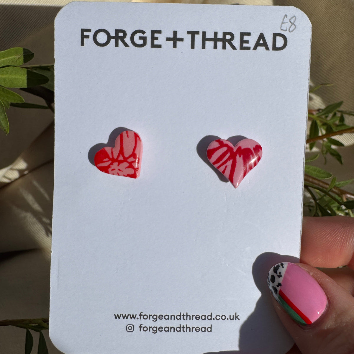 A pair of red and white patterned heart stud earrings displayed on a white Forge+Thread card, held by a hand with pink and green nail art against a beige fabric background with green leaves.