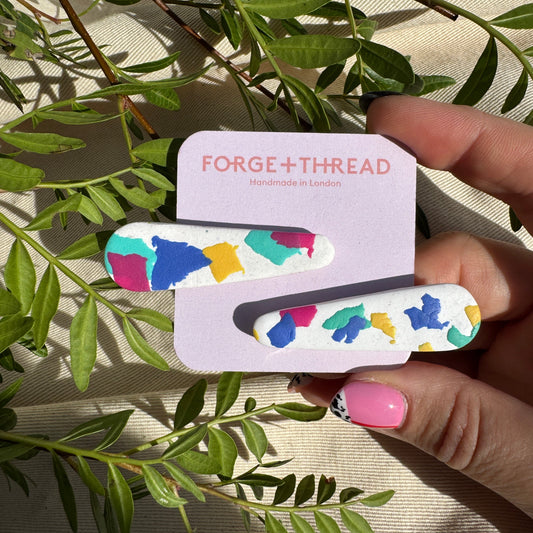 A hand holds a lavender card displaying two matching hair clips with a colorful abstract pattern. The card is branded 'FORGE+THREAD'.