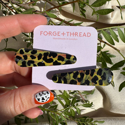 A hand holds a white display card featuring two green, yellow, and black leopard-print hair clips. The card is branded 'FORGE+THREAD' and is styled with green leaves and cream fabric in the background.