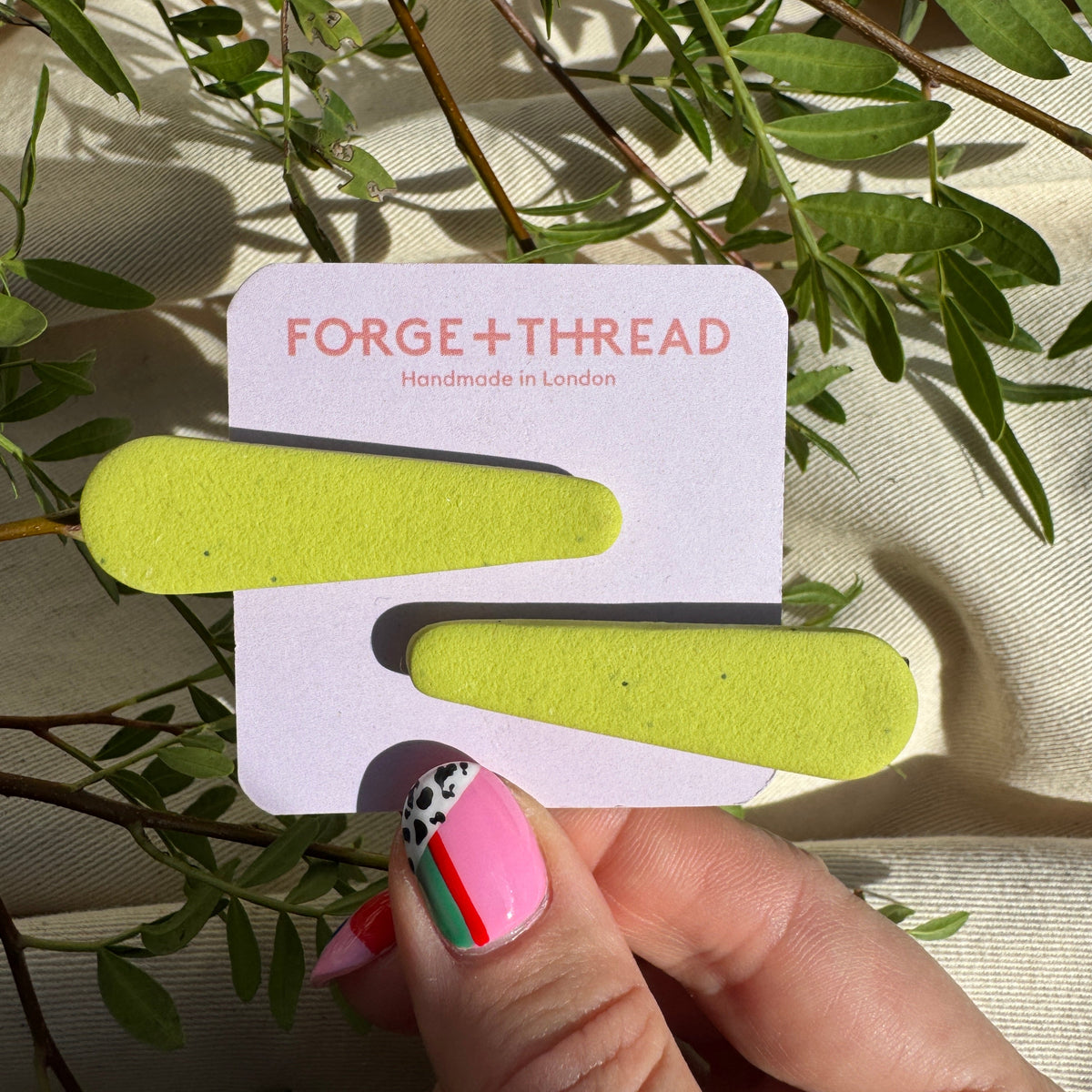 A hand with decorated pink nails holds a white card displaying two bright yellow-green hair claw clips against a background of green leaves and beige fabric. The card is branded 'FORGE+THREAD'.