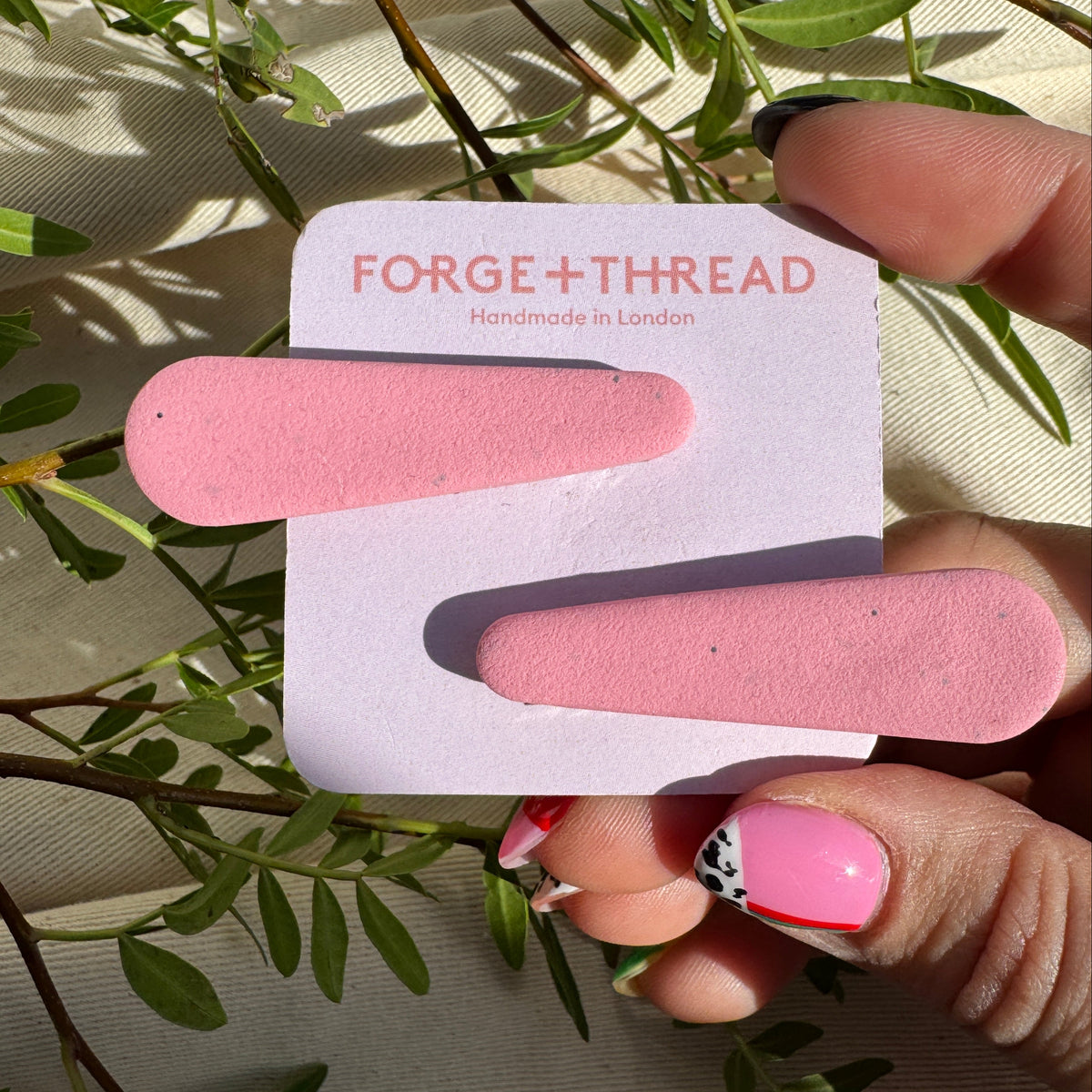 A hand holds a white display card featuring two pink, elongated nail files or emery boards. The card is printed with the brand name 'FORGE+THREAD' and 'Handmade in London'.
