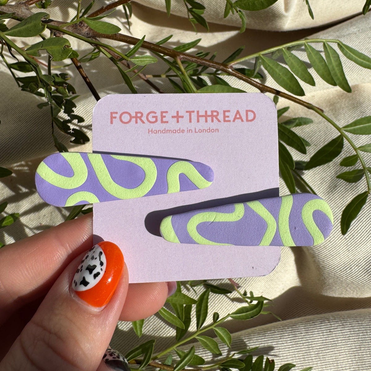 A hand with orange and black nail art holds a lavender card displaying two matching hair clips with a purple and neon green wavy pattern. The card is branded 'FORGE+THREAD Handmade in London' and is styled with green leaves on a beige fabric background.