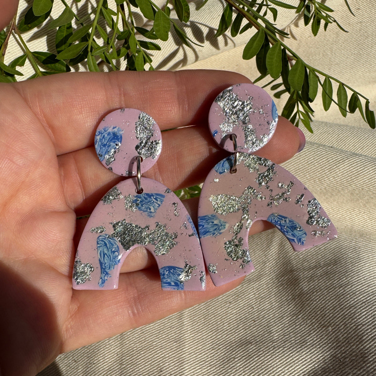 A pair of handmade statement earrings held in a hand. The earrings feature a dusty pink base with blue floral patterns and silver glitter accents, with a matching round stud on top.
