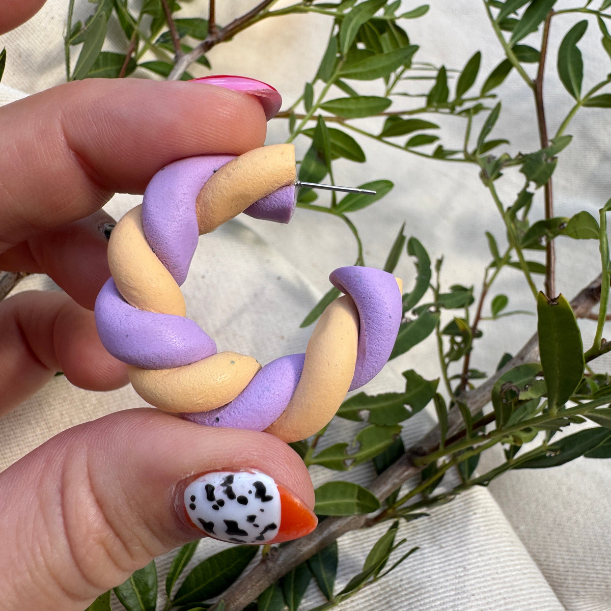 A close-up of a hand holding a twisted, braided ring made of lavender purple and pale peach segments, with green leaves and a beige fabric background.